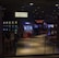 A dimly lit casino with several rows of slot machines, some displaying bright graphics and lights. There are a few people scattered throughout the area, engaging with the machines. The floor is reflective and polished, adding to the ambiance, while the ceiling is dotted with numerous small lights.