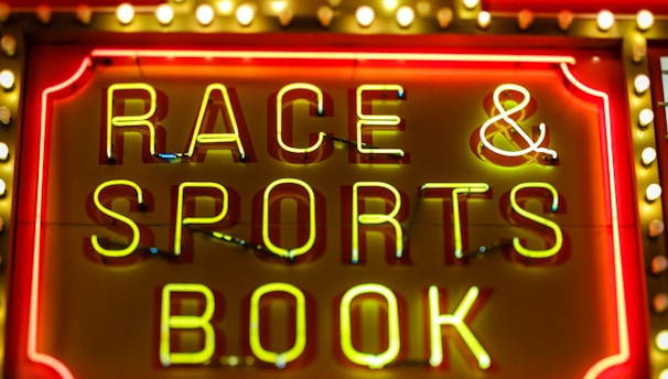 Bright neon lights on a marquee sign display the words 'RACE & SPORTS BOOK'. The sign is surrounded by rows of yellow and white lights against a red background, evoking a vibrant, energetic atmosphere typically associated with casinos or entertainment venues.