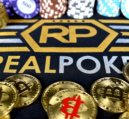 Stacks of poker chips in various colors are arranged in the background, while in the foreground, there are numerous golden coins featuring the Bitcoin symbol. The text 'REALPOKER' is prominently displayed in gold and silver lettering on a dark background.