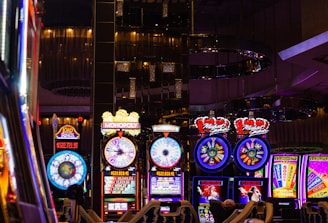 A lively casino scene featuring a row of bright, colorful slot machines with themes like Monopoly, Michael Jackson, and Lord of the Rings. The gaming machines display a variety of flashing lights and digital screens, creating an energetic and exciting atmosphere. Reflections of the lights are visible in glossy surfaces above the machines.