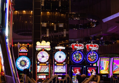 A lively casino scene featuring a row of bright, colorful slot machines with themes like Monopoly, Michael Jackson, and Lord of the Rings. The gaming machines display a variety of flashing lights and digital screens, creating an energetic and exciting atmosphere. Reflections of the lights are visible in glossy surfaces above the machines.