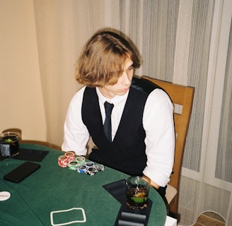 A person with shoulder-length hair is sitting at a poker table. They are wearing a white shirt, black vest, and tie, with an array of poker chips in front of them. There are two glasses with drinks on the table, and a smartphone is placed nearby. The background consists of vertical striped curtains.
